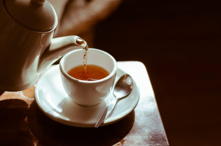 Une théière verse du thé chaud dans une tasse blanche posée sur une soucoupe, avec une cuillère posée à côté. Un éclairage chaleureux crée une ambiance chaleureuse sur la table en bois.