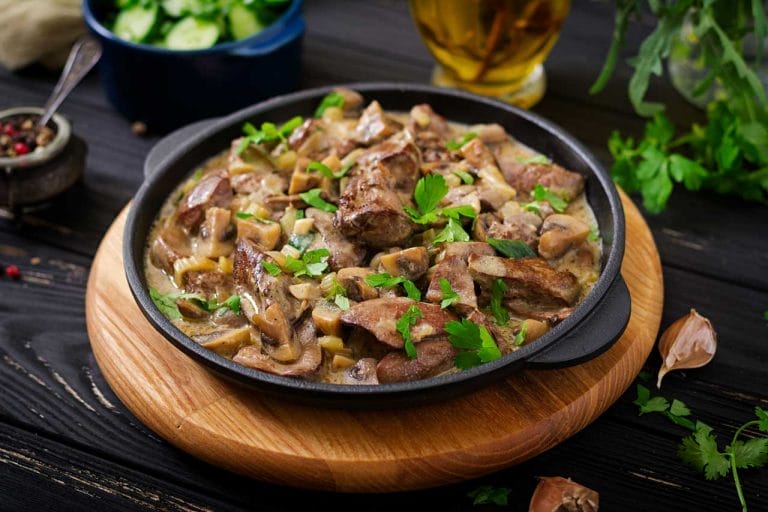 Une poêle remplie d'un plat crémeux de viande cuite et de champignons, garni de persil frais. La poêle repose sur une planche de bois, entourée d'ingrédients frais et d'un bol de concombres tranchés en arrière-plan.