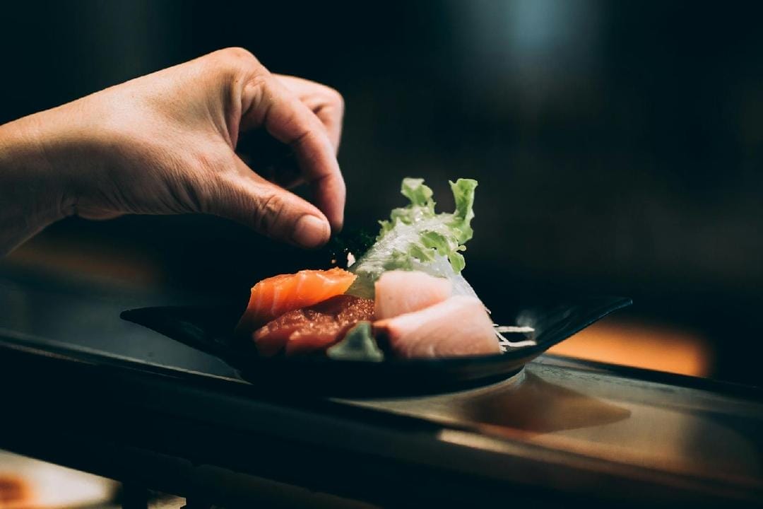 Une main dépose une garniture sur une assiette de sashimi, composée de tranches de saumon et de poisson blanc, avec de la laitue sur une assiette sombre. La scène est capturée dans un éclairage doux et chaleureux, mettant en valeur l'apparence fraîche et invitante du plat.