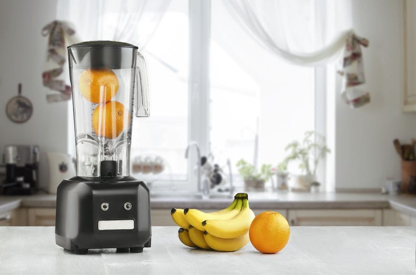 Un blender noir contenant deux oranges entières est posé sur un comptoir de cuisine. À côté se trouvent un régime de bananes et une orange supplémentaire. L'arrière-plan présente une fenêtre lumineuse avec des rideaux transparents et des éléments de décoration de cuisine.