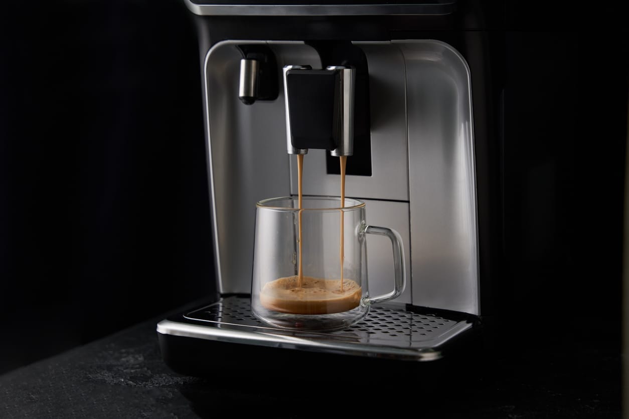 Une tasse en verre transparent est posée sous une machine à café. Deux jets de café s'y déversent, créant une couche de crème sur le café. L'arrière-plan sombre met en valeur le processus d'infusion.