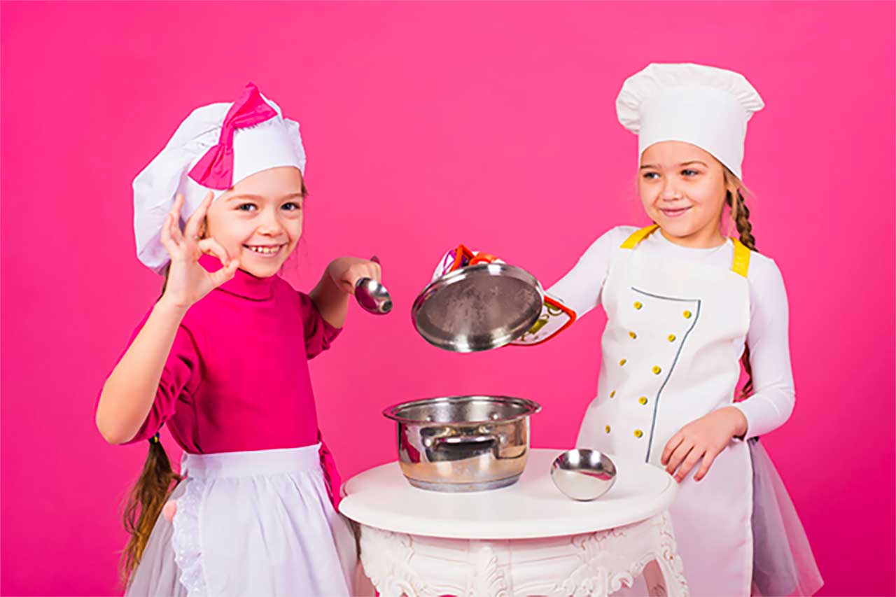 Deux jeunes filles portant des toques et des tabliers se tiennent à une petite table blanche avec une casserole, en souriant. L'une d'elles tient un couvercle et fait un geste de la main pour dire "OK", tandis que l'autre se tient à côté d'elle. L'arrière-plan est rose vif.