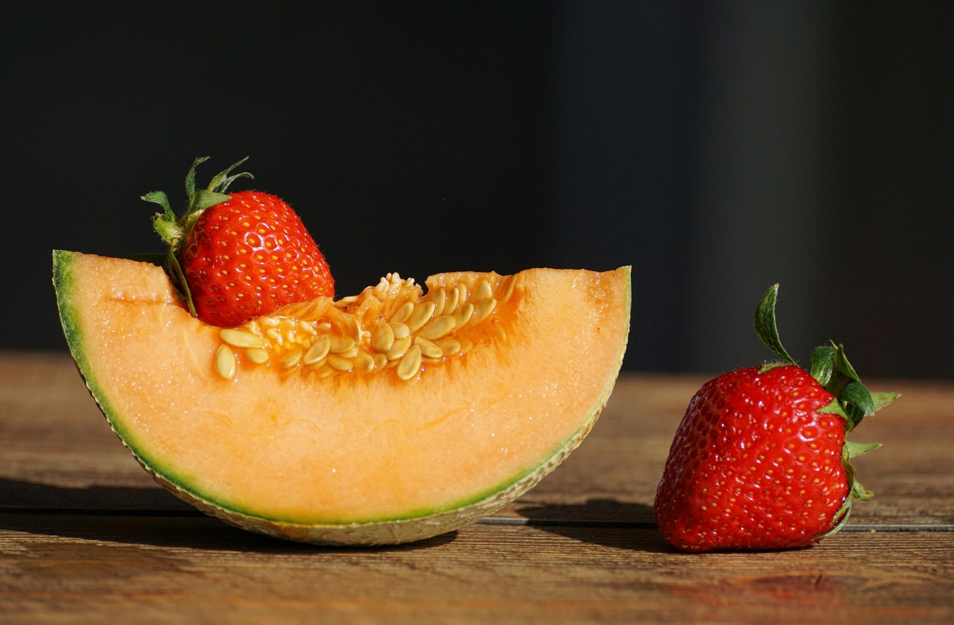 Une tranche de cantaloup avec des graines est posée sur une surface en bois, une fraise repose sur le melon et une autre fraise est placée à côté.