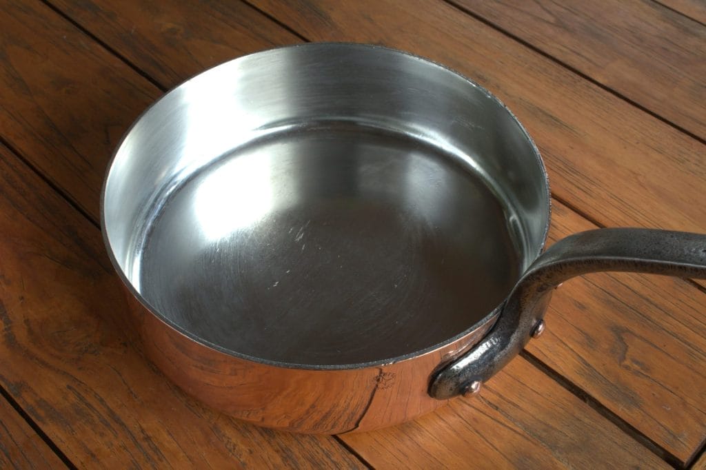 Une casserole en métal vide et brillante, dotée d'un long manche, repose sur une surface en bois. On peut y voir l'intérieur réfléchissant et l'extérieur cuivré.