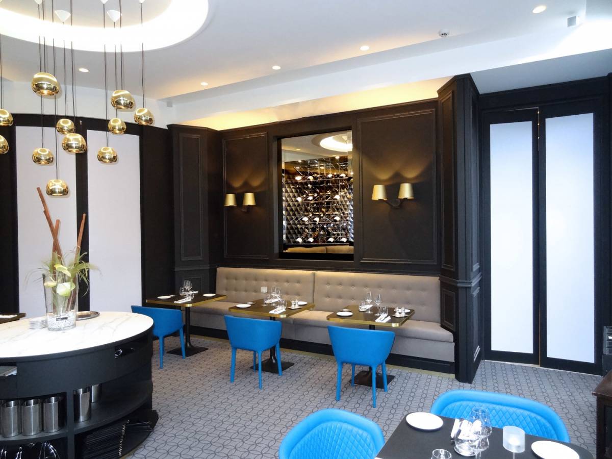 Intérieur d'un restaurant moderne avec des murs recouverts de panneaux sombres, un grand miroir, des banquettes grises rembourrées, des chaises bleues, des tables bien rangées, des éclairages décoratifs et un vase avec de hautes branches sur un chariot de service rond.