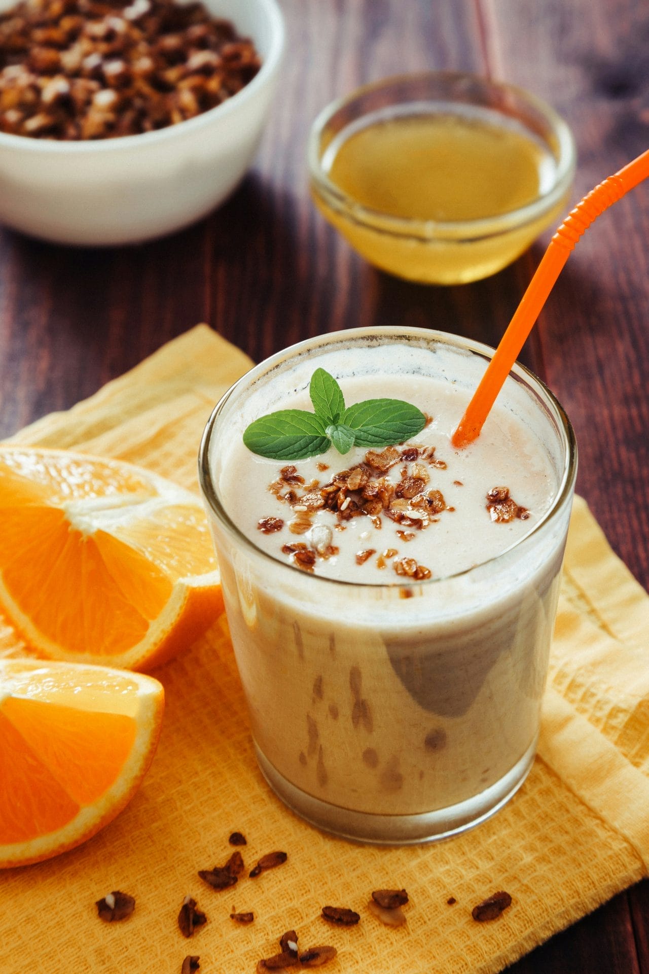 Un verre de smoothie garni de granola et de menthe, avec une paille orange, est posé sur une serviette jaune à côté de tranches d'orange. À l'arrière-plan, des bols de granola et de miel sont posés sur une table en bois.