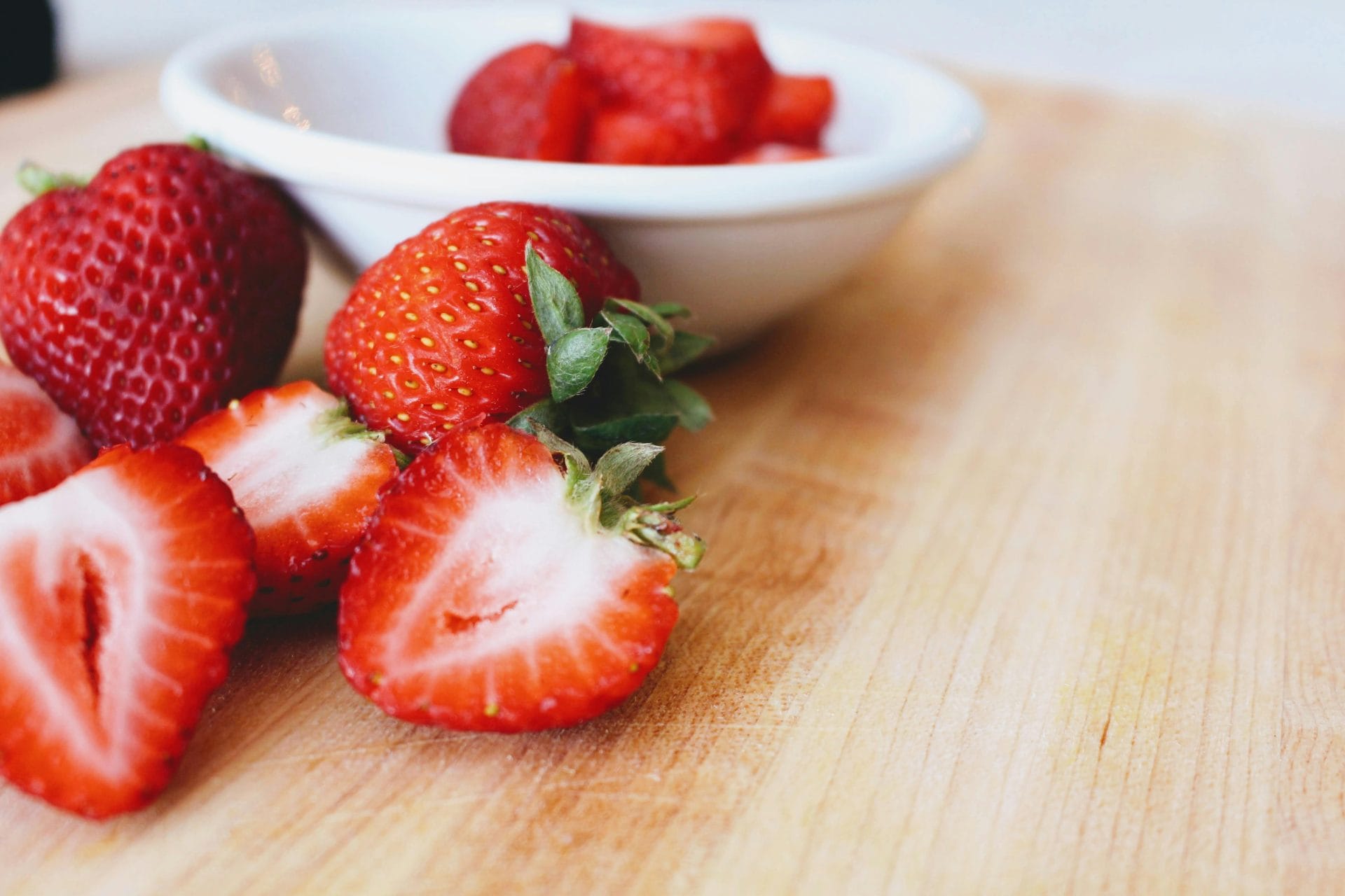 Des fraises fraîches, certaines entières et d'autres coupées en deux, sont disposées sur une surface en bois clair. Un bol blanc contenant d'autres fraises tranchées se trouve à l'arrière-plan.