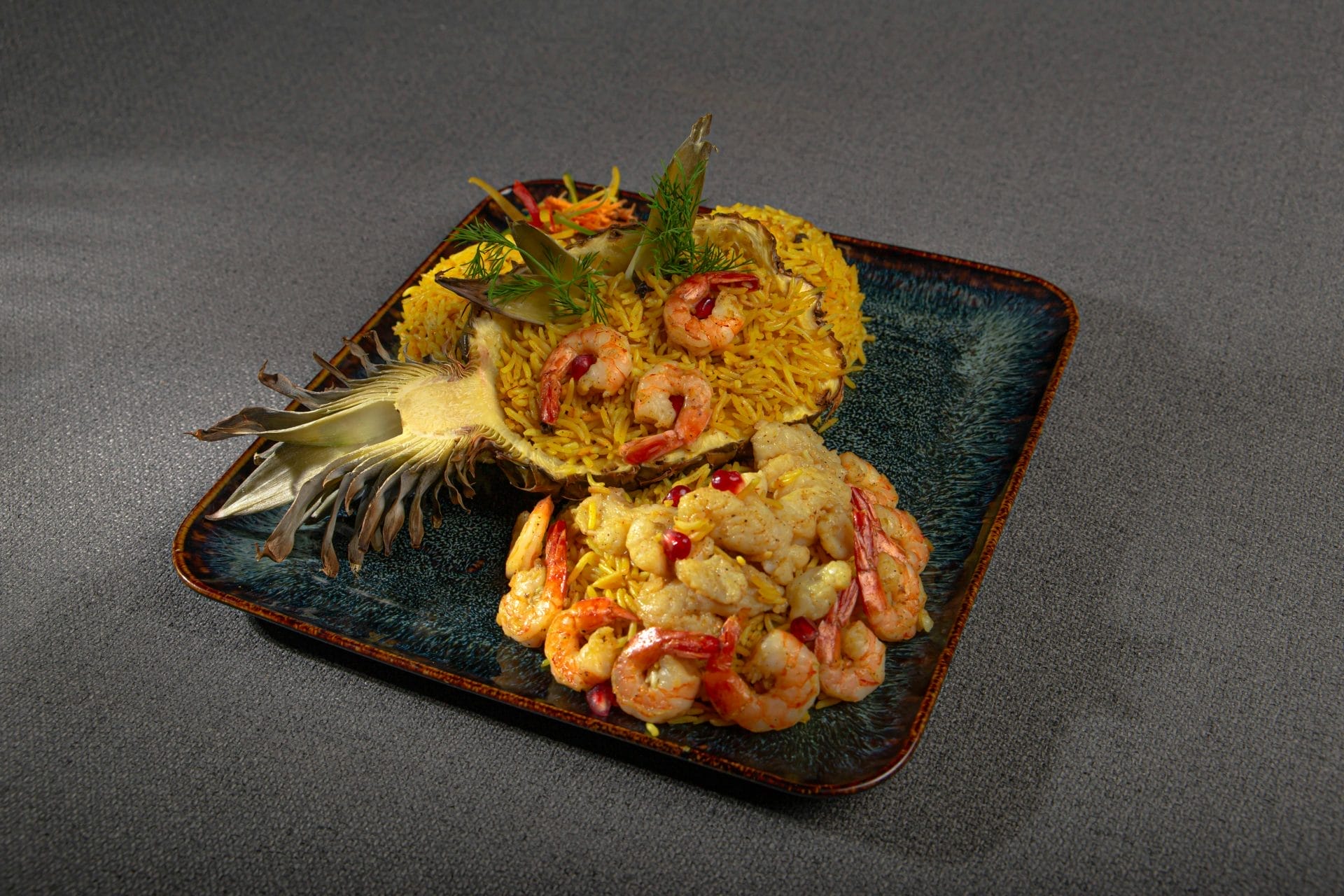 Assiette carrée contenant du riz jaune garni de crevettes, servi avec un sauté de crevettes et de légumes. Un ananas décore le plat, qui est présenté sur une surface grise texturée.