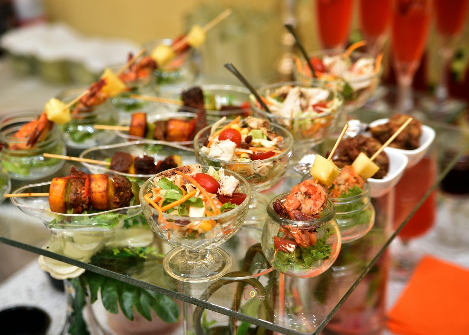 Diverses entrées dans de petits bols et pots en verre, dont des brochettes de crevettes, des légumes grillés et de la salade, disposées sur un plateau en verre avec des verres de boissons roses à l'arrière-plan.