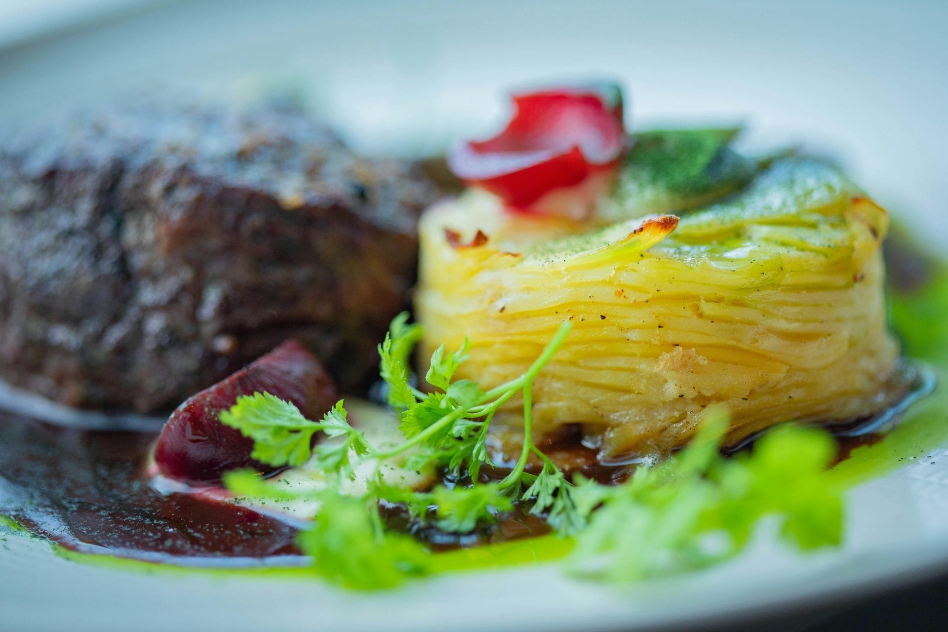 Gros plan sur un plat gastronomique composé d'une pile de pommes de terre coupées en fines tranches, agrémentées d'herbes et de sauce, à côté d'une portion de viande cuite et de fleurs comestibles sur une assiette blanche.