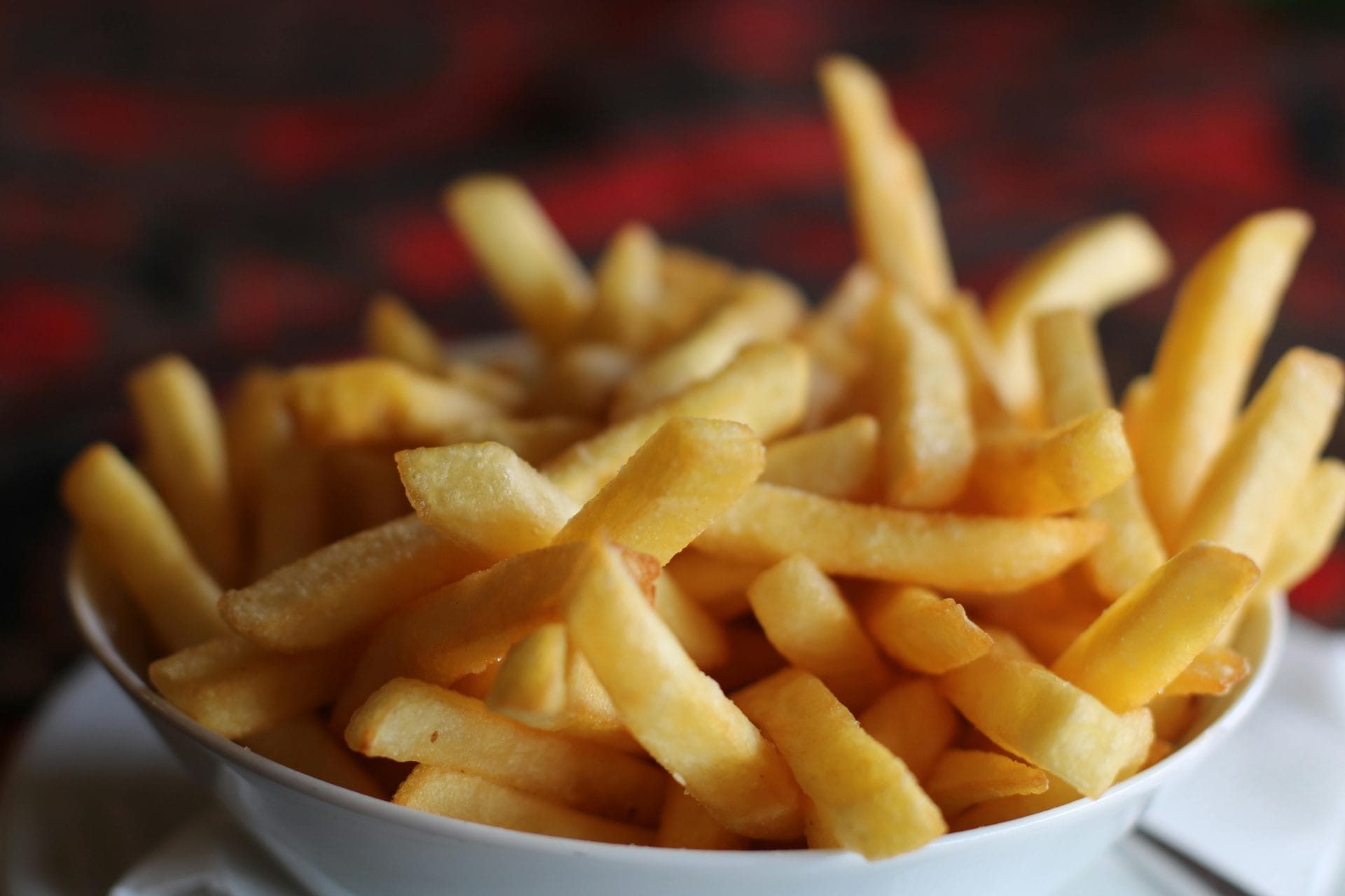 Gros plan d'un bol blanc rempli de frites dorées et croustillantes, sur un fond flou rouge foncé et noir.