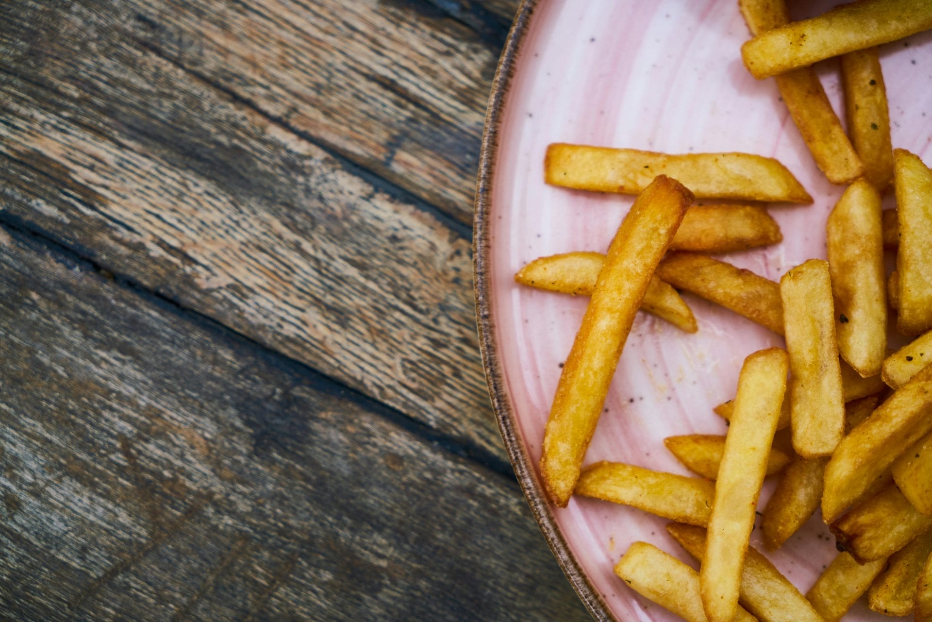 Frites dorées sur une assiette rose, posée sur une table rustique en bois. Quelques frites sont éparpillées, l'assiette n'étant que partiellement visible dans le coin supérieur droit de l'image.