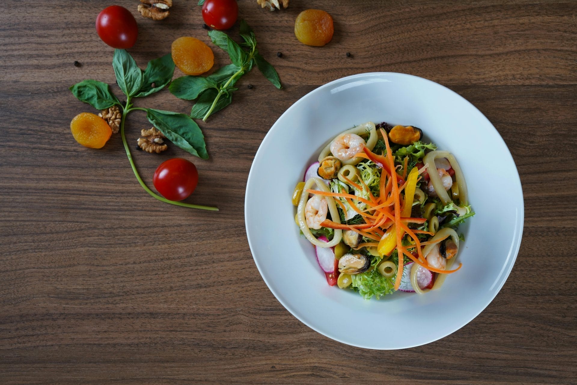 Une assiette de salade de fruits de mer avec des crevettes, des olives et des carottes râpées, garnie de verdure. Sur la table en bois se trouvent des tomates cerises, des feuilles de basilic, des abricots secs et des noix.