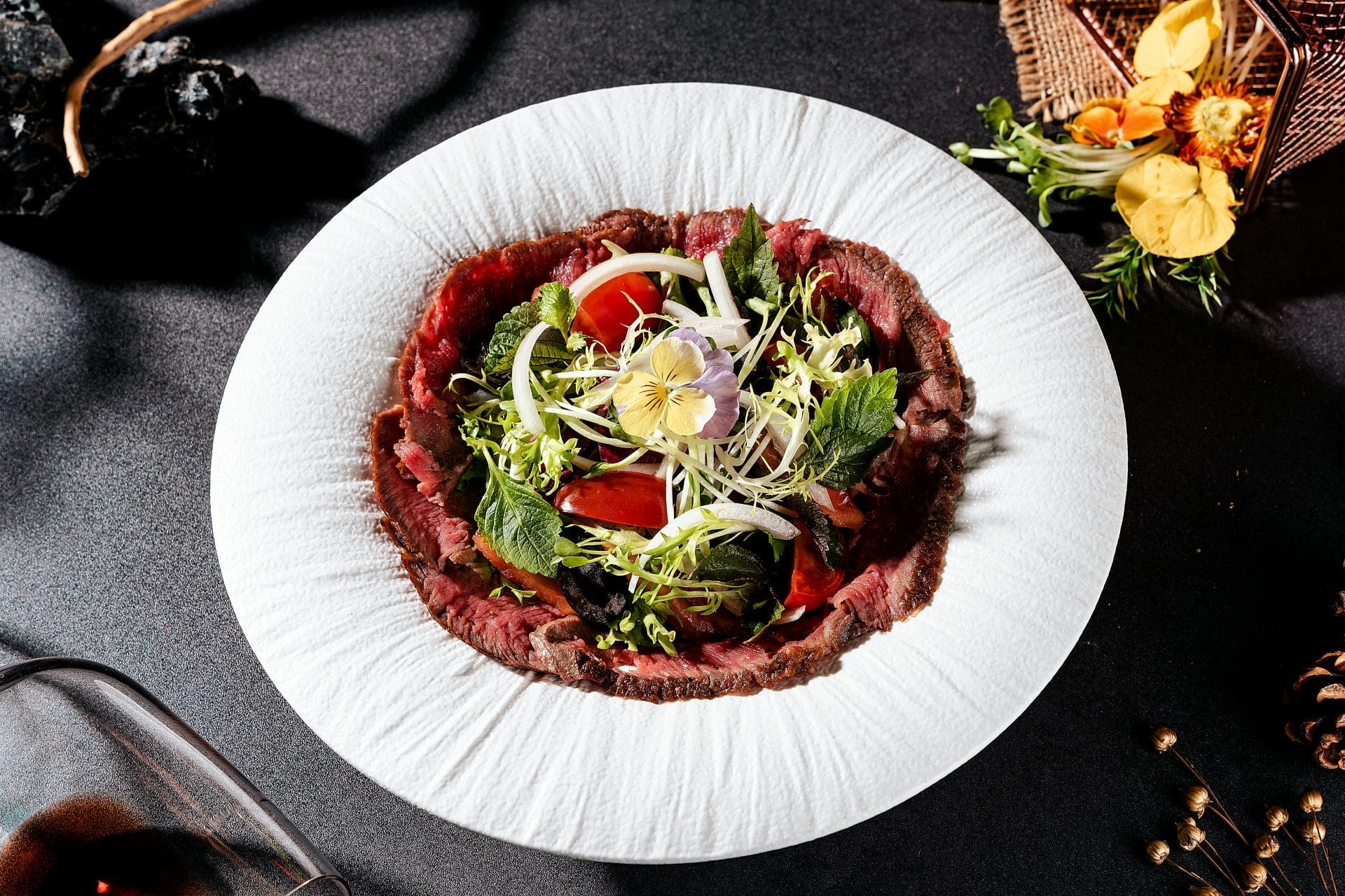 Une assiette blanche contenant un carpaccio de bœuf finement tranché, garni d'un mélange de verdure, de pousses, d'herbes et de fleurs comestibles, le tout disposé avec art sur une table sombre.