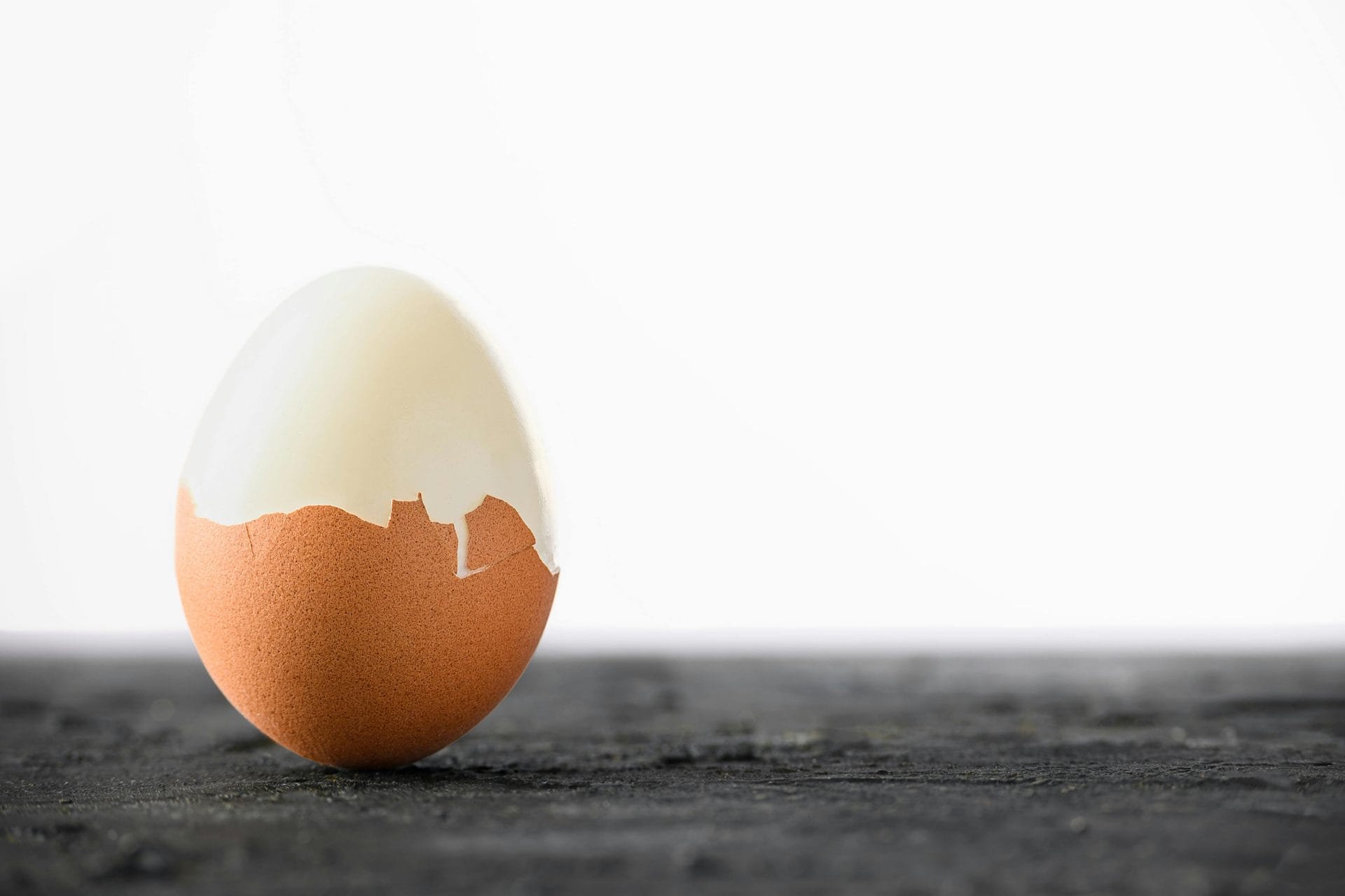 Un œuf dur, dont la coquille a été partiellement écalée, est posé à la verticale sur une surface sombre, sur un fond blanc uni.