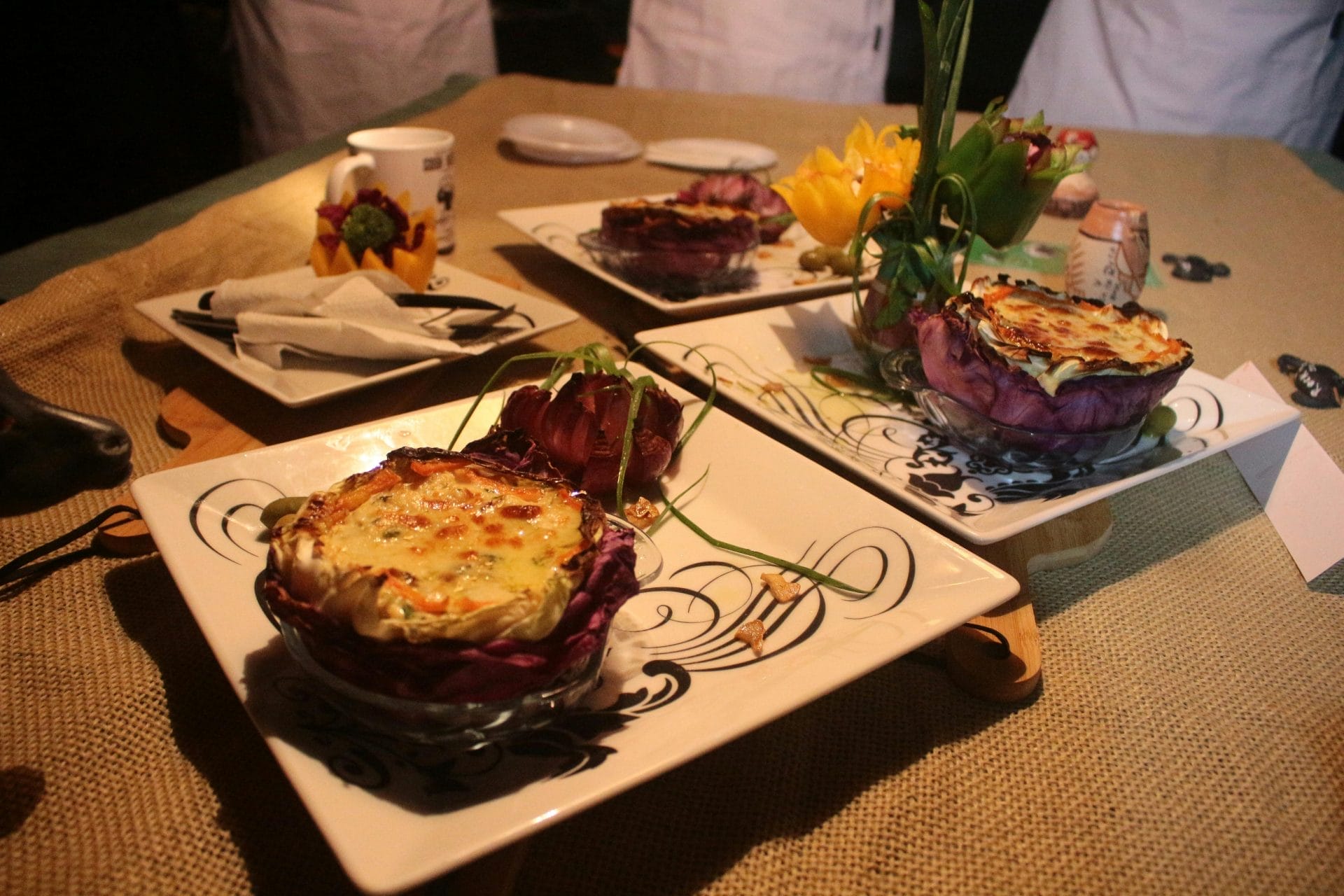 Plats gastronomiques servis dans des bols à choux violets surmontés de fromage fondu, disposés sur des assiettes carrées décoratives, sur une table recouverte de toile de jute, avec couverts, gobelets et serviettes à proximité.