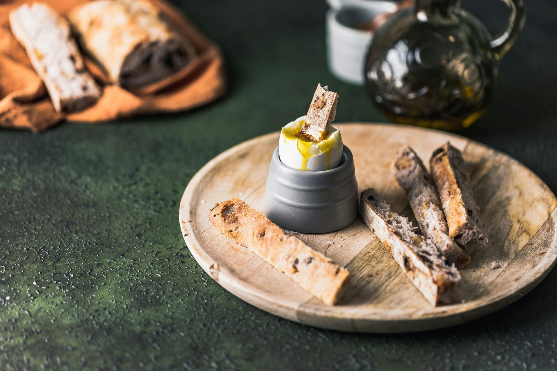 Bâtonnets de pain tranchés sur une assiette en bois, dont un morceau est trempé dans de l'huile d'olive dans un petit bol. Une miche de pain et une bouteille d'huile d'olive se trouvent à l'arrière-plan.