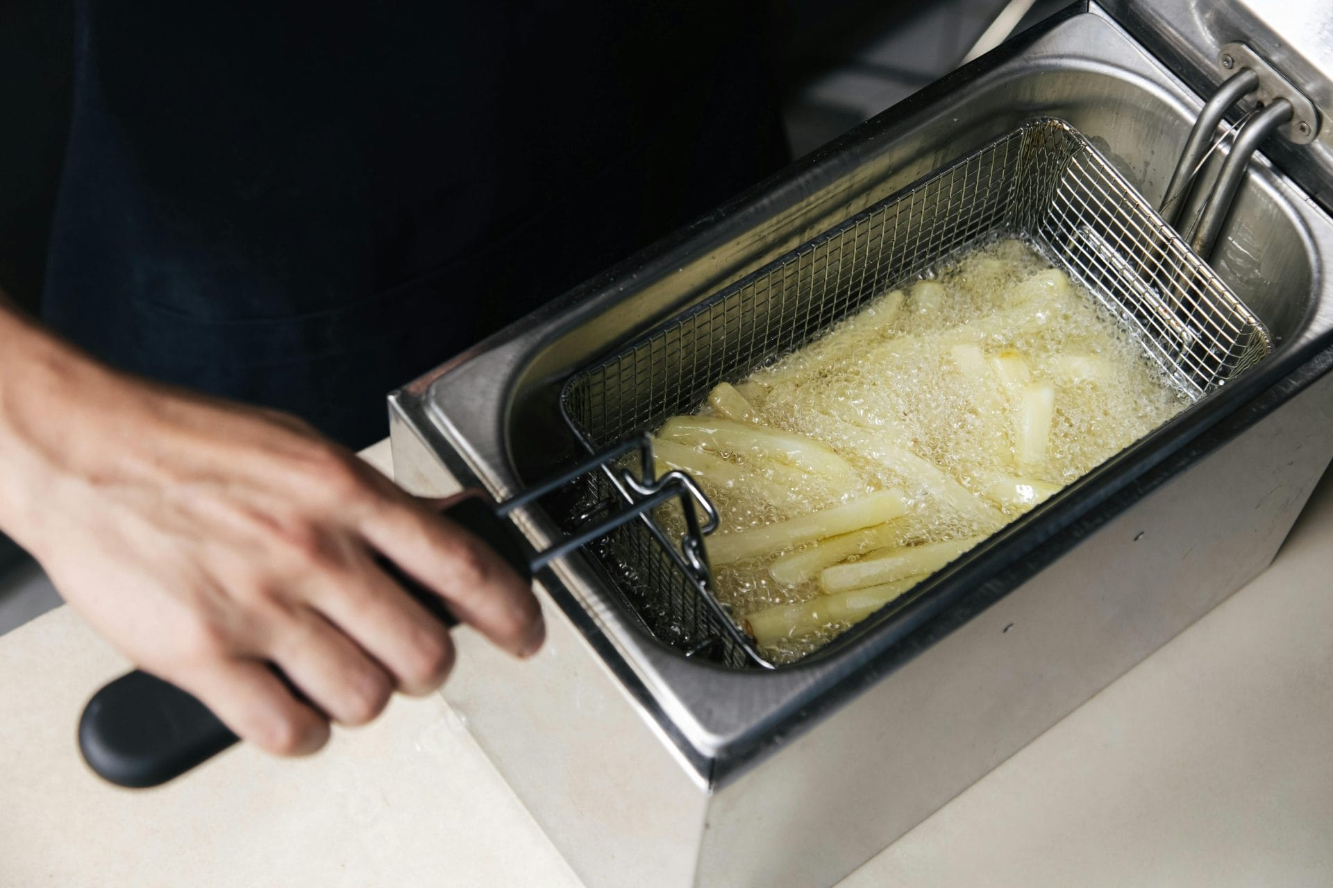 Une personne tient la poignée d'un panier métallique contenant des frites de pommes de terre en train d'être frites dans de l'huile chaude à l'intérieur d'une friteuse commerciale.