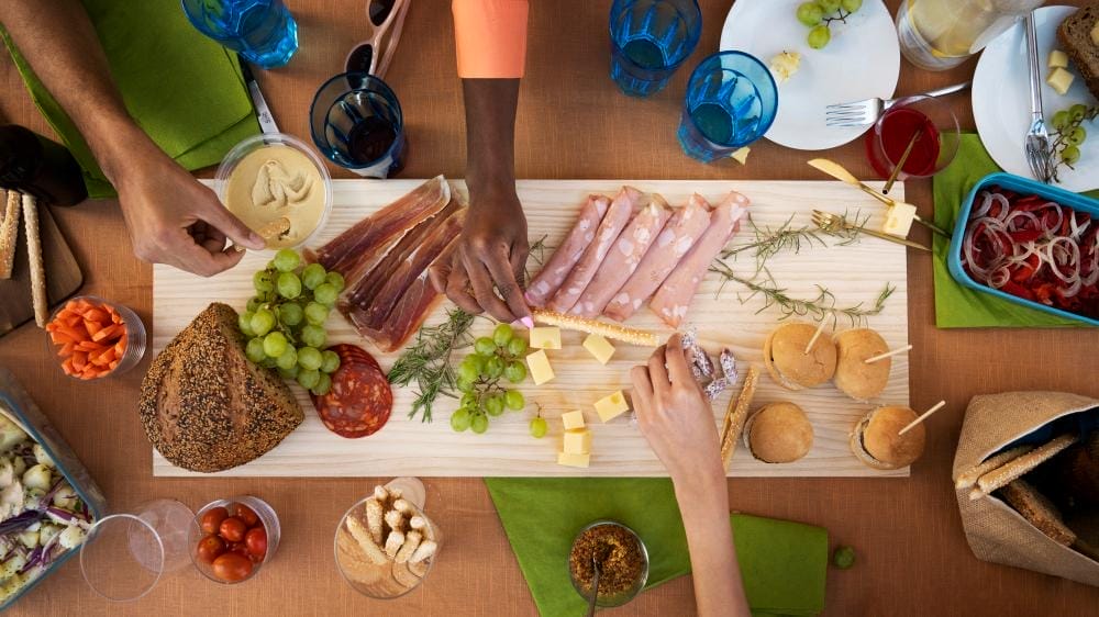 Vue de haut en bas d'une table sur laquelle se trouve une planche en bois sur laquelle sont disposés des viandes, des cubes de fromage, des raisins, du pain et des sauces. Plusieurs mains tendent la main pour attraper de la nourriture, avec des assiettes, des boissons et des petits sandwichs autour de la planche.