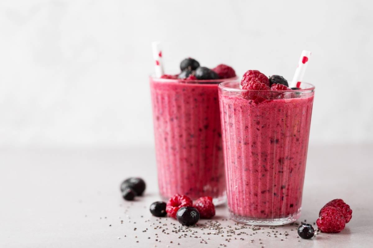 Deux verres remplis de smoothie aux baies rose vif, garni de framboises et de myrtilles. Chaque verre est muni d'une paille blanche avec des cœurs rouges. Des baies supplémentaires et des graines de chia sont éparpillées sur la surface claire.