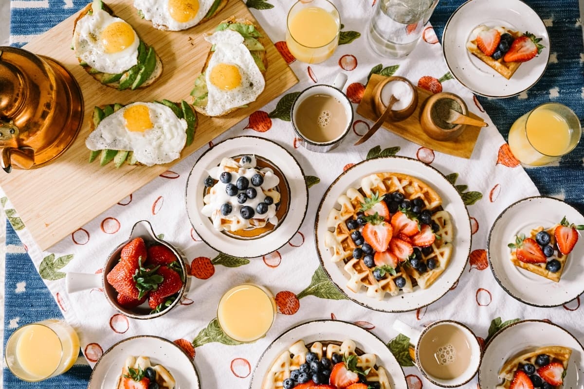 Une table de petit-déjeuner composée de gaufres garnies de baies, de toasts d'avocat avec des œufs au plat, de bols de fraises, de verres de jus d'orange, d'une théière en cuivre et d'une nappe blanche avec un motif de fruits rouge et vert.